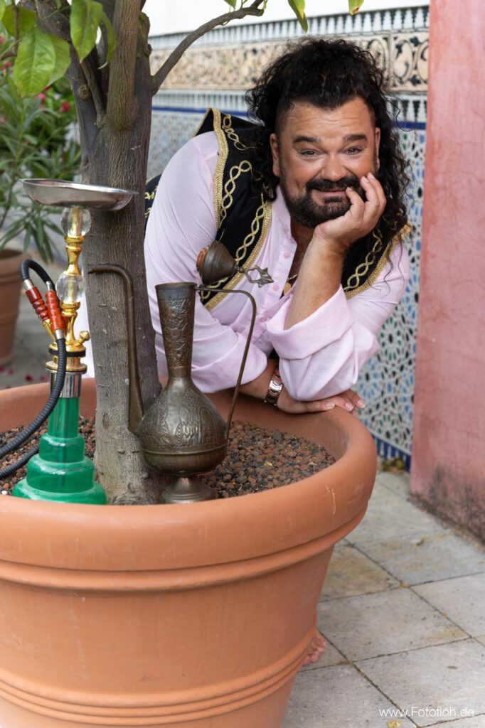 🇬🇧 Mustafa Roshan in traditional Persian-Afghan clothing enjoying a shisha in the Gardens of the World in Berlin, surrounded by palm trees and oriental garden elements. 🇩🇪 Mustafa Roshan im traditionellen persisch-afghanischen Outfit mit einer Shisha in den Gärten der Welt in Berlin, umgeben von Palmen und orientalischen Gartenelementen.